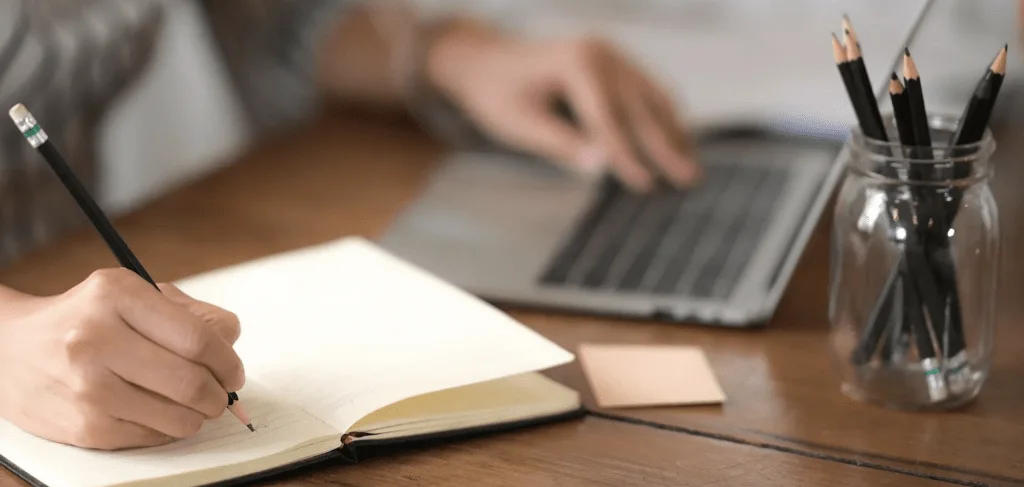 Person writing in a notebook beside an open laptop and a jar of pencils.